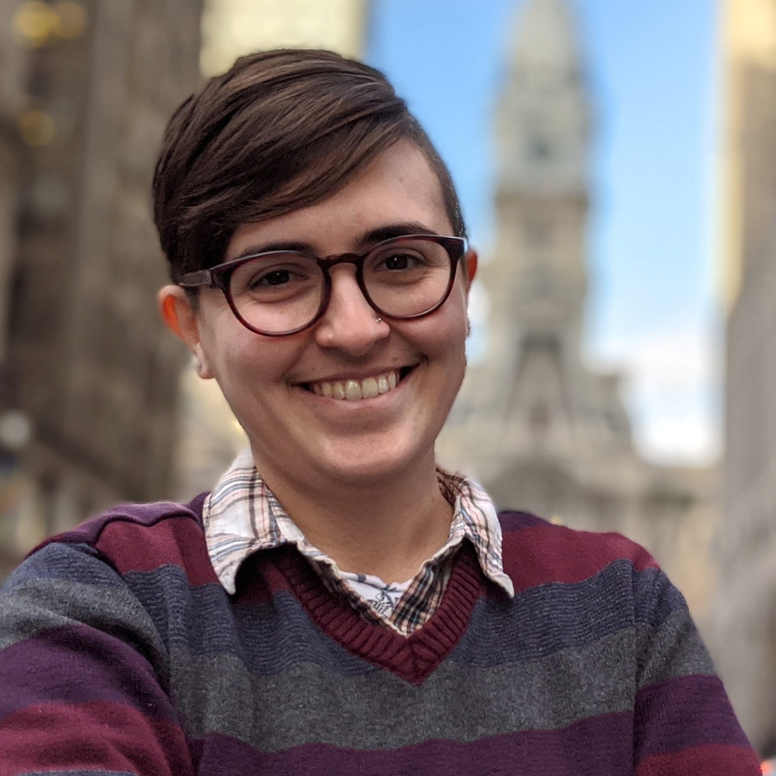 Portrait of a light-skinned person with short, tidy light-brown hair and glasses, with a beaming smile wearing a striped shirt with collar with a skyscraper in the distant background.