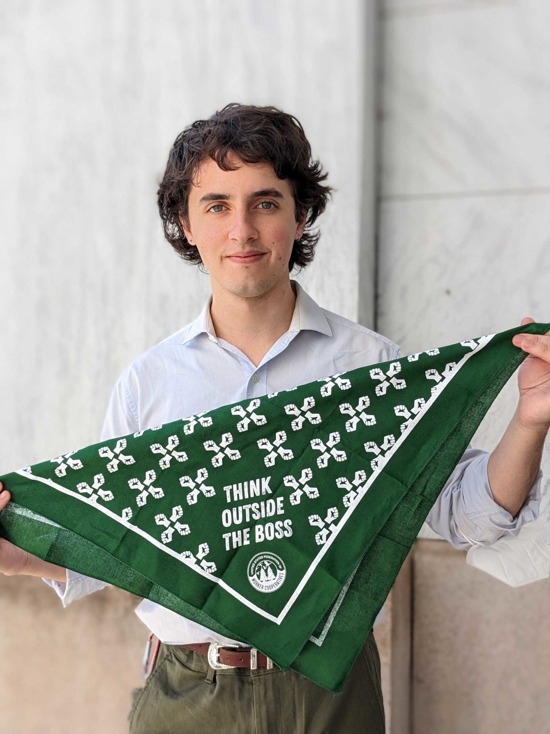 A picture of a person with a white collared shirt dark brown hair and blue eyes holding a dark green bandana that says "think outside the Boss"