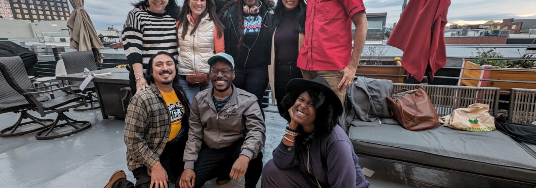 Group of 9 people of different races and genders posing together and smiling joyfully on a rooftop patio overlooking a city skyline.