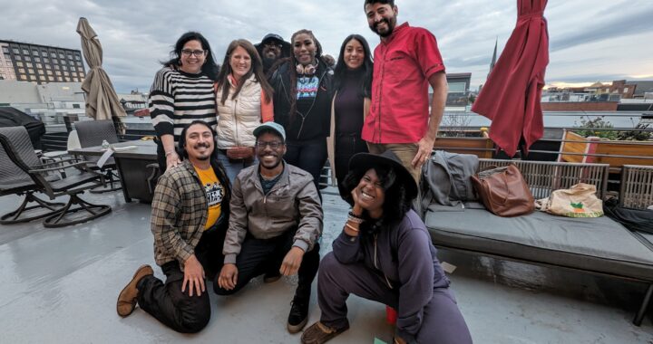 Group of 9 people of different races and genders posing together and smiling joyfully on a rooftop patio overlooking a city skyline.