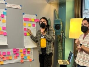 Tall, slender person with brown skin and long, straight black hair wearing a flannel shirt and black slacks and a covid mask motions to a large white paper with lots of colorful sticky notes on it.
