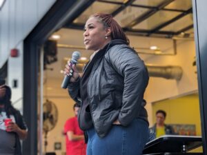 Black woman with crimson braids and a black hoodie under a grey rain jacket speaks confidently on the mic in an outdoor patio.