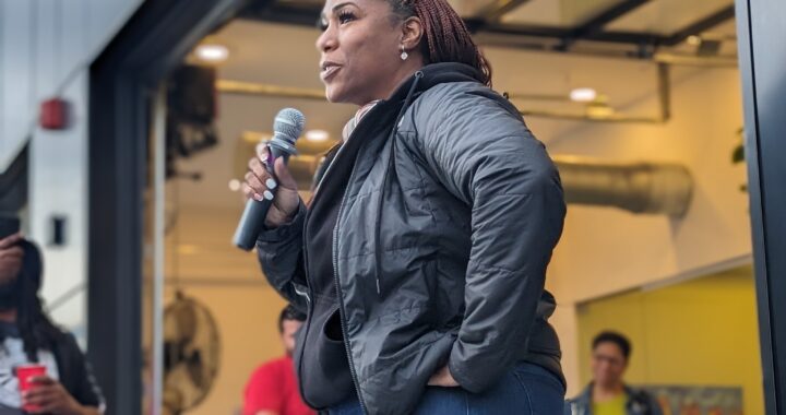 Black woman with crimson braids and a black hoodie under a grey rain jacket speaks confidently on the mic in an outdoor patio.