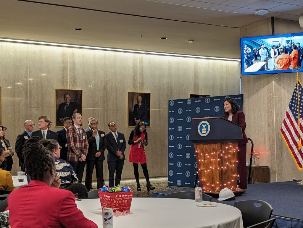 Acting Secretary Julie Su [Asian woman wearing a burgundy suit] speaking at a Department of Labor event, December 2023. A crowd of people listens to the Acting Secretary speaking on stage at a podium decorated with holiday lights.