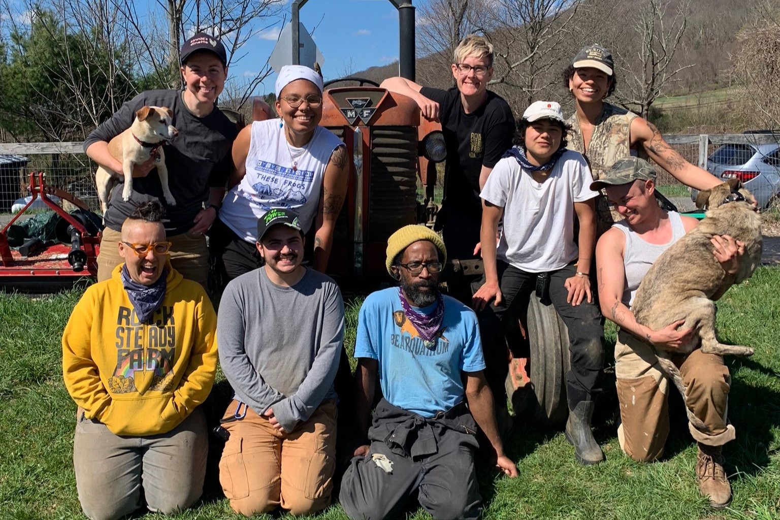 A photo of nine people kneeling and standing outside on a tractor with two dogs. The relatively young people are diverse in race color and gender expression and smile to the camera."