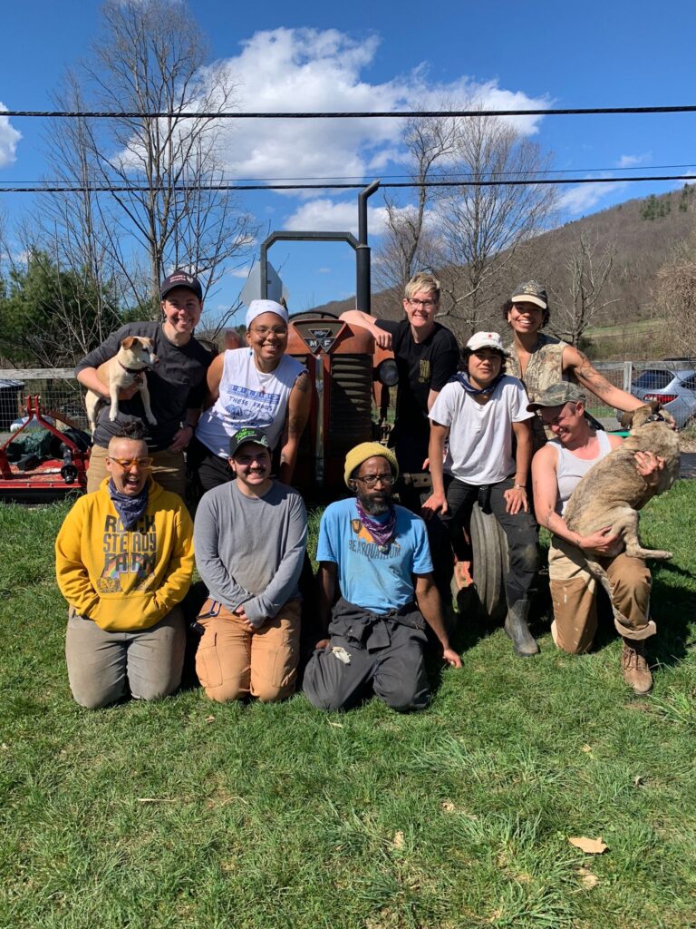 A photo of nine people kneeling and standing outside on a tractor with two dogs. The relatively young people are diverse in race color and gender expression and smile to the camera.