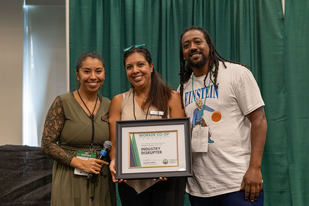 Portrait of three people in a stage smiling while posing shoulder-to-shoulder. The person on the right has dark brown skin, black hair worn in braids that fall to the chest, and a mustache and beard. He wears a white t-shirt that says "Einstein" with an illustration of the scientist playing basket ball. The person in the middle has shoulder-length dark brown hair, light brown skin, wears a pair of sunglasses on top of their head, a light brown blouse and holds a framed certificate that says "Industry disrupter." The person on the left has dark brown hair pulled back into a ponytail, light brown skin, wears an olive-colored dress with lace sleeves and holds a microphone.