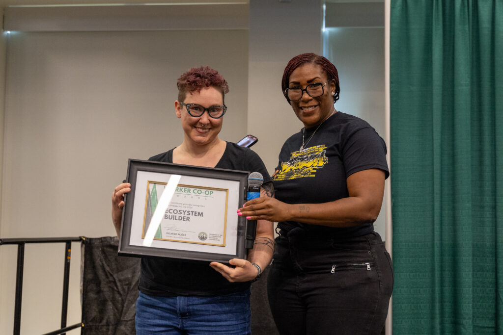 A photo of two people standing on a stage. Person on the left has short pink hair and wears eye glasses and holds a framed certificate that reads ecosystem builder. Person on the right has long red braids, wears eyeglasses and smiles holding the framed certificate and a microphone.