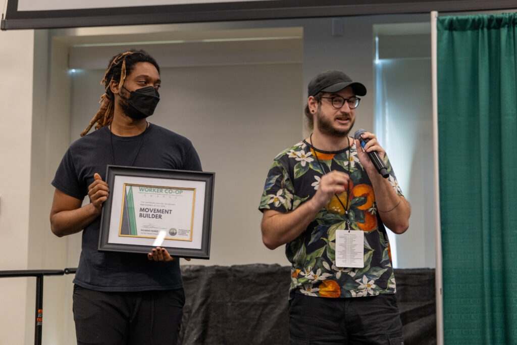 A photo of two people on a stage. Person on the left has long bleached locs and wears and a surgical face mask and holds a framed award certificate with text that reads worker co-op awards movement builder. Person on the right wears a black hat and a floral shirt and a lanyard and speaks into a microphone.