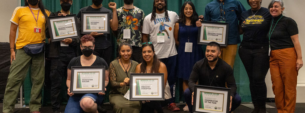 People of different races and genders on a stage stand shoulder to shoulder and smile joyfully while displaying framed certificates.