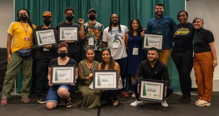 People of different races and genders on a stage stand shoulder to shoulder and smile joyfully while displaying framed certificates.