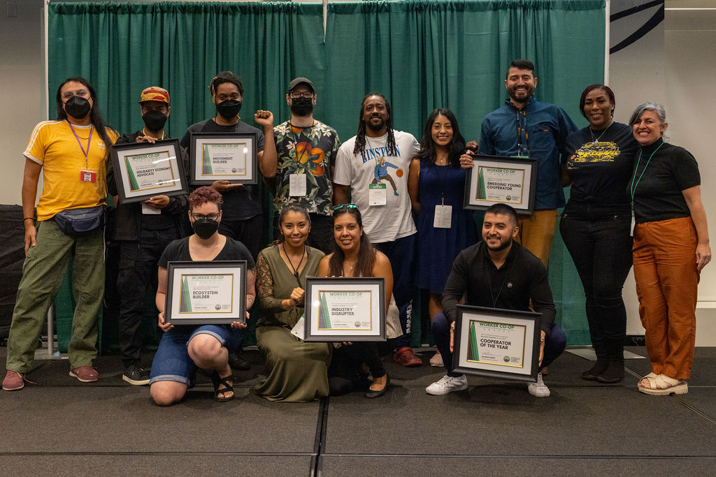 People of different races and genders on a stage stand shoulder to shoulder and smile joyfully while displaying framed certificates.