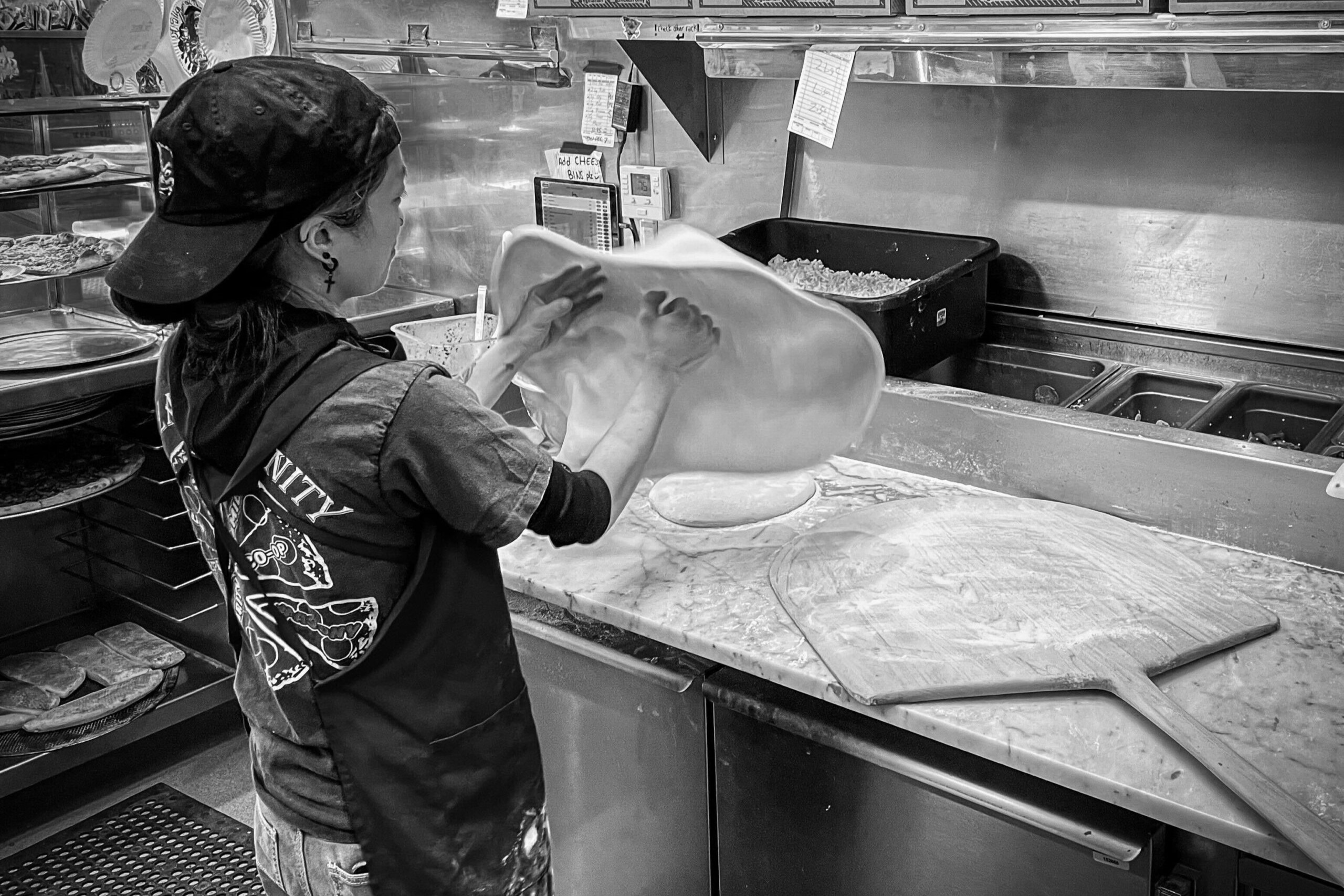 A photo of a short person wearing a backwards baseball cap and apron tossing a circular piece of pizza dough in the air and spinning it. Pizza boxes and other ingredients can be seen in the background."