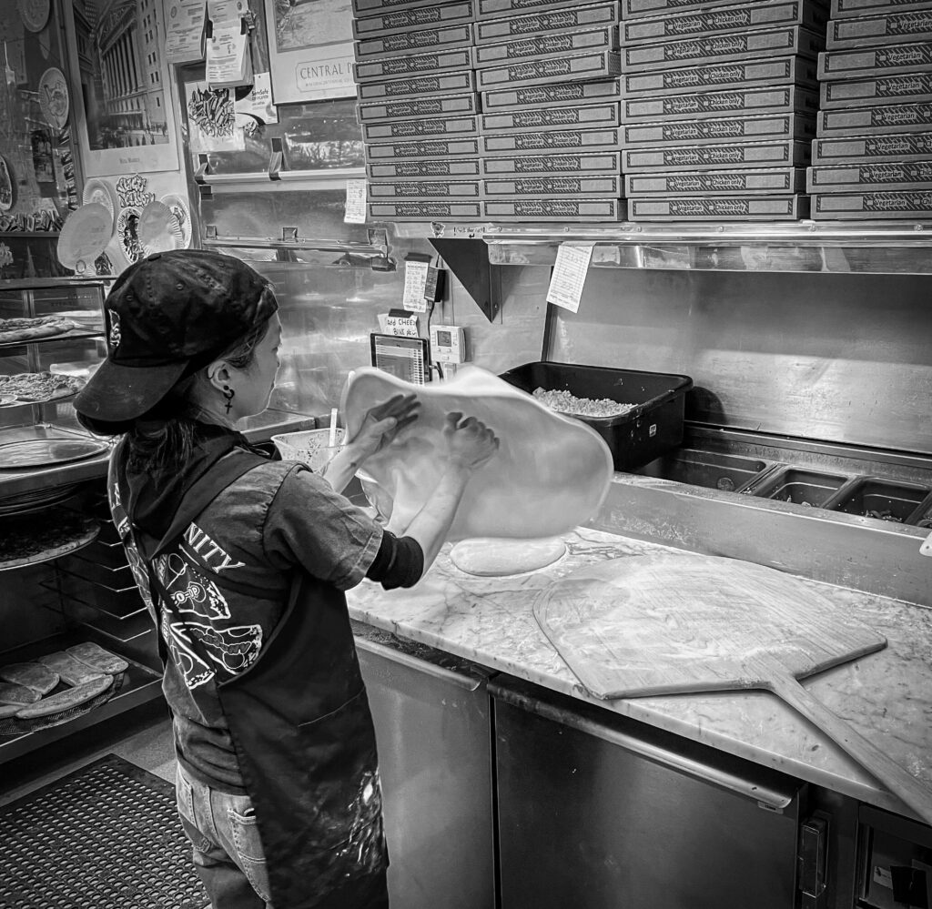 A photo of a short person wearing a backwards baseball cap and apron tossing a circular piece of pizza dough in the air and spinning it. Pizza boxes and other ingredients can be seen in the background.