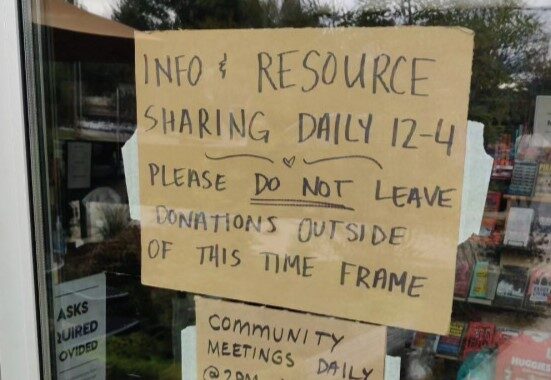 makeshift cardboard sign in storefront window that says "info and resource sharing daily 12 to 4. Community meetings daily at 2 pm around back"