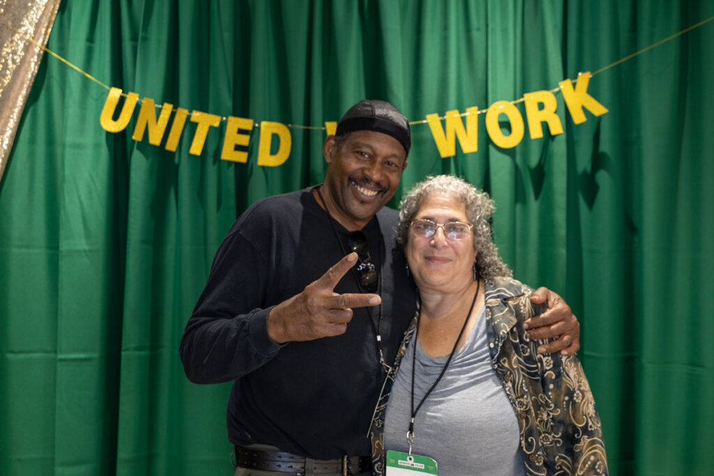 Two people posing together in front of a green curtain and a garland hung with gold letters that read "United .... Work". The person on the left has black skin, a mustache, and short hair covered in a durag. They are making a peace sign with their right hand and grinning broadly while their left arm is wrapped around a shorter person wearing glasses with long curly gray hair and light skin.