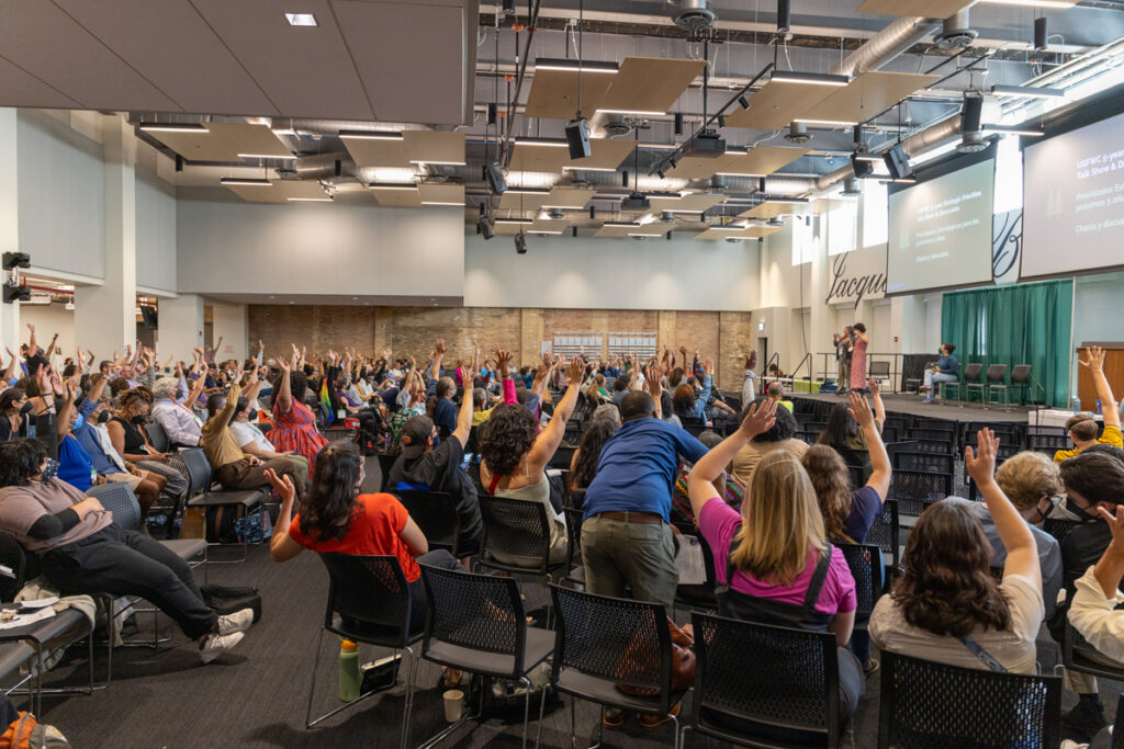 A large conference hall filled with people sitting with hands raised.