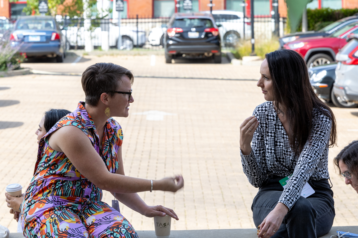 Two individuals sit outside a city building aside a parking lot engaged in a deep conversation, one leans in toward the other gesturing while the other peers on intently.