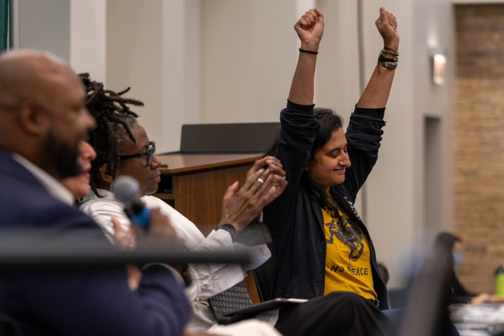 Person with brown skin and shoulder length black hair raises arms triumphantly while person with black skin and thin braids done up in a bun looks on and applauds.