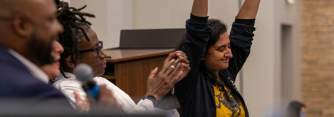 Person with brown skin and shoulder length black hair raises arms triumphantly while person with black skin and thin braids done up in a bun looks on and applauds.