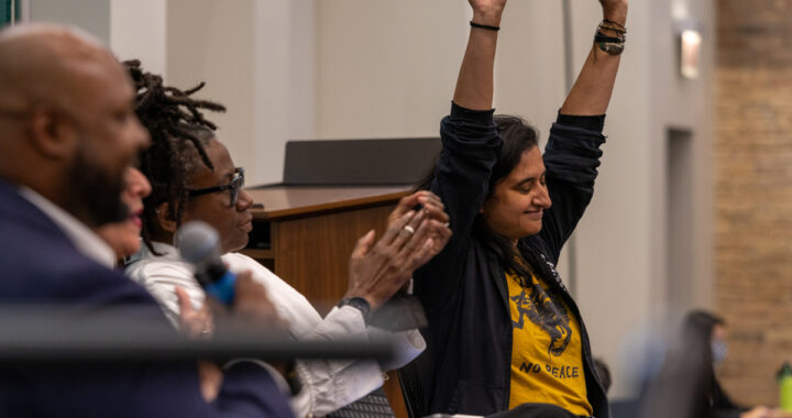 Person with brown skin and shoulder length black hair raises arms triumphantly while person with black skin and thin braids done up in a bun looks on and applauds.