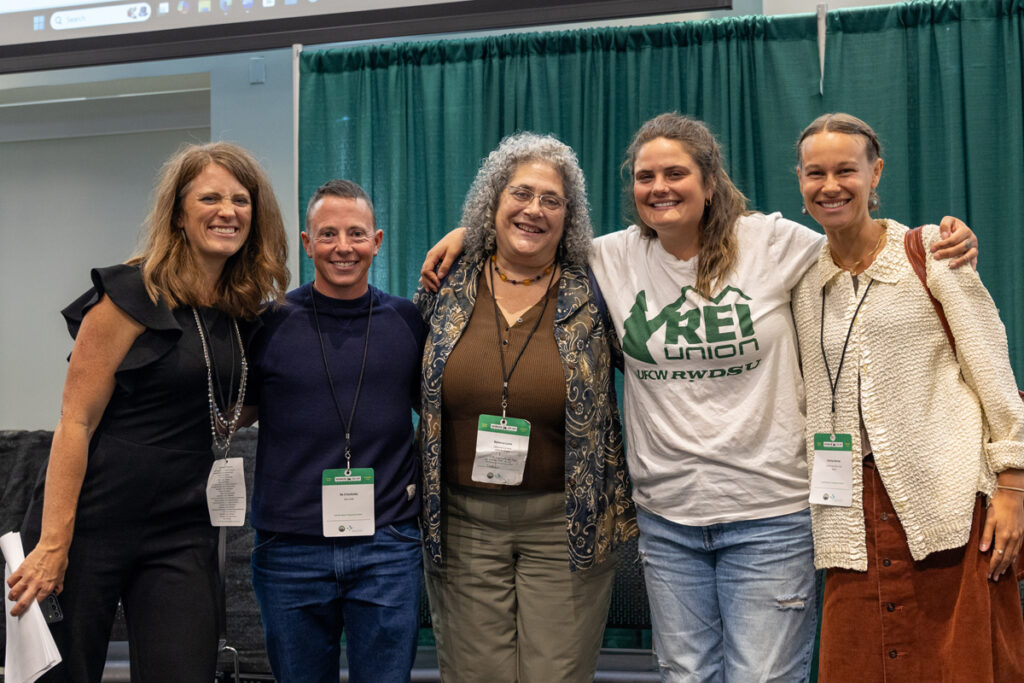 Five people with light skin standing together and smiling at the camera with their arms around each other's shoulders. One of the people's shirts reads "REI union".