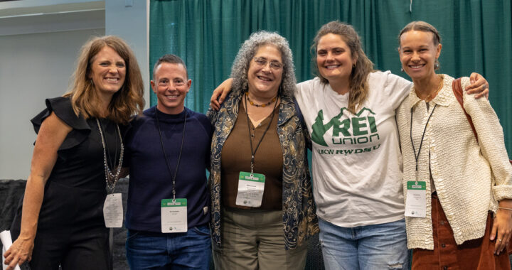 Five people with light skin standing together and smiling at the camera with their arms around each other's shoulders. One of the people's shirts reads "REI union".