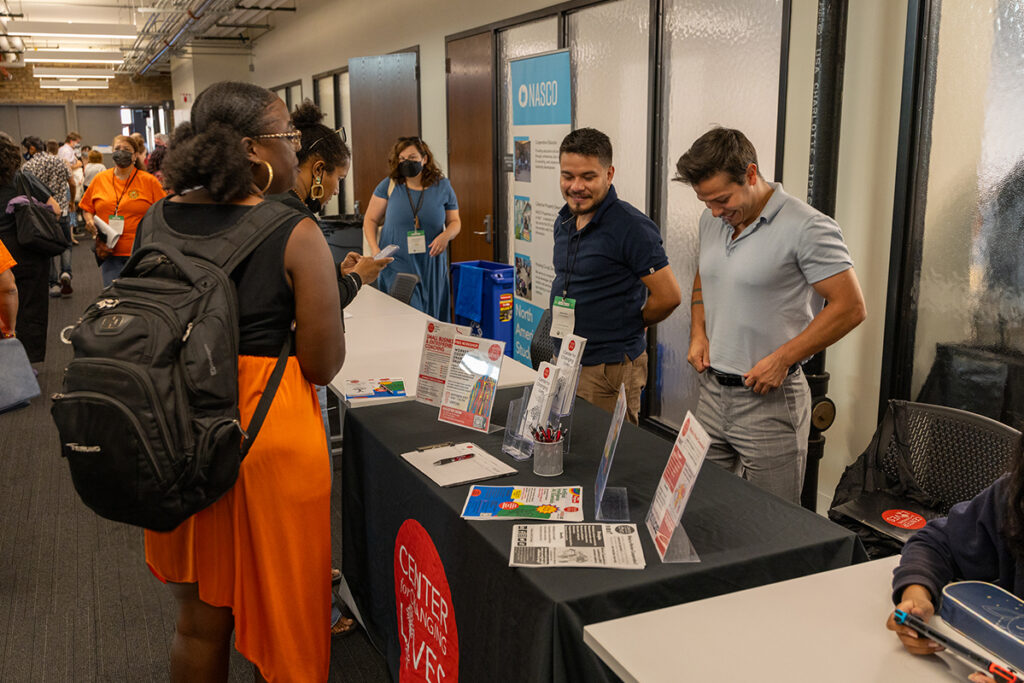 Person with black skin and short hair in pigtails talking to two people in polo shirts standing behind a table. The table is covered in a black tablecloth with a large red logo on the front, and on top of it are signs, fliers, handouts, and a signup clipboard.