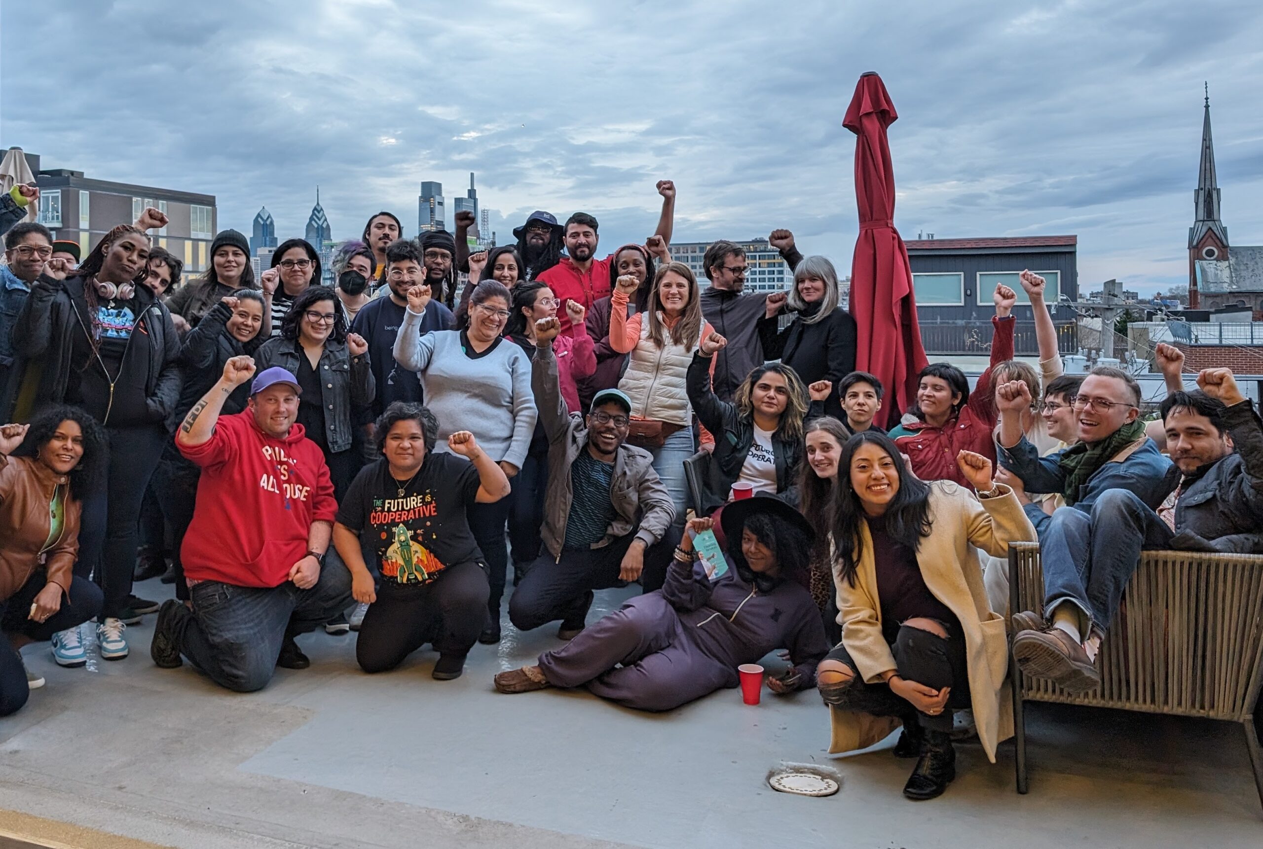 Group of people of different genders, races, and ages posing with raised fists and broad smiles on a rooftop patio overlooking a cityscape.